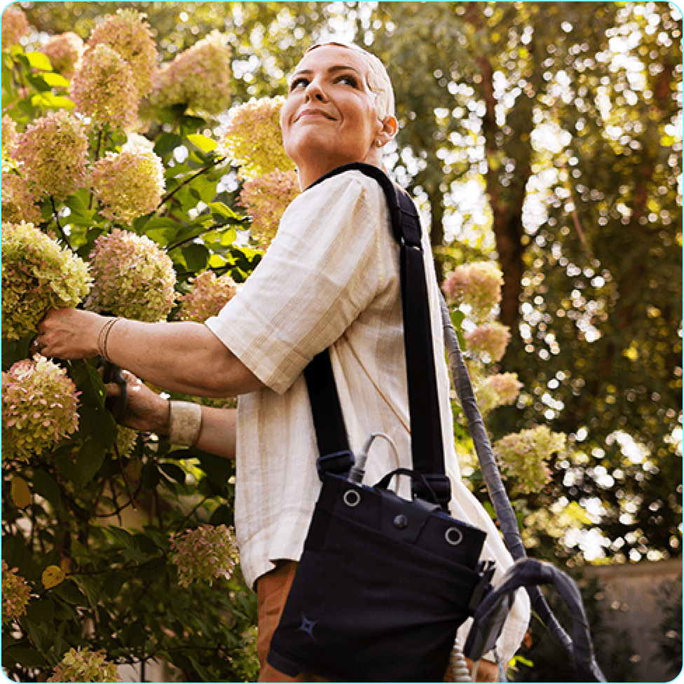 Ryann outdoors in a garden setting while using Optune Gio with the device shoulder carrying case.